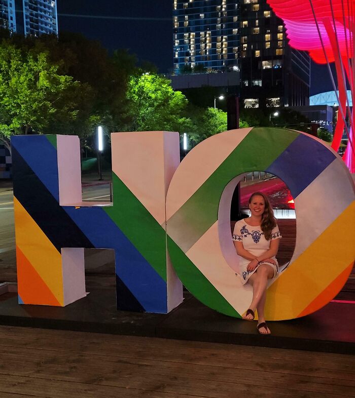 Woman sitting cheerfully inside a colorful "HO" installation at night, surrounded by city lights and trees.