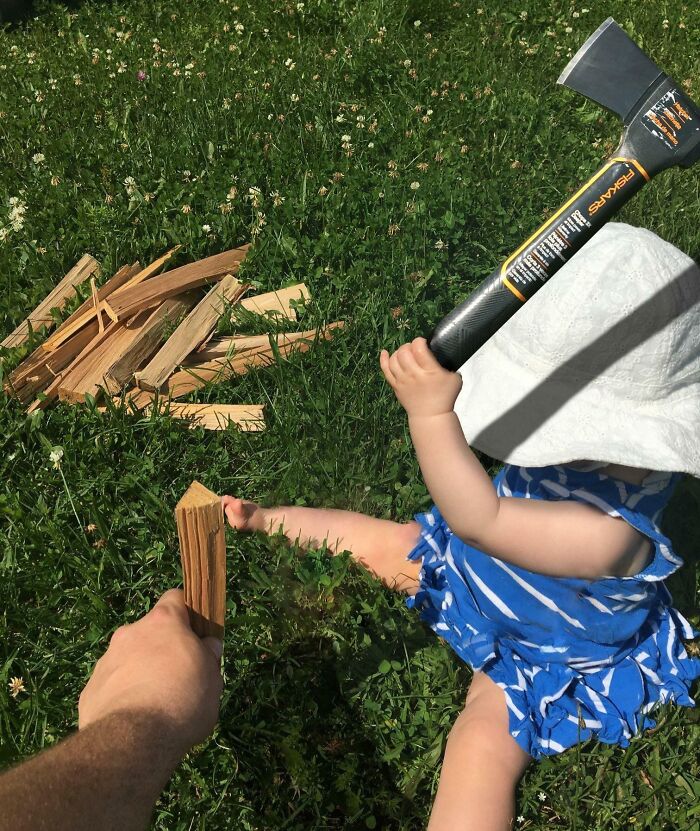 Toddler holding an axe sitting on grass, adult hand offers wood; humorous scene with a partner's playful twist.