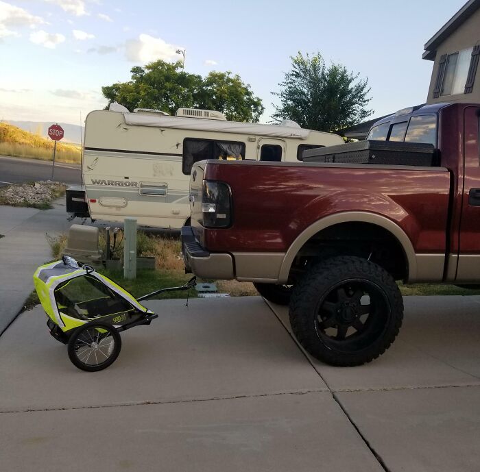 Truck humor with a red pickup towing a tiny bike trailer in a driveway.