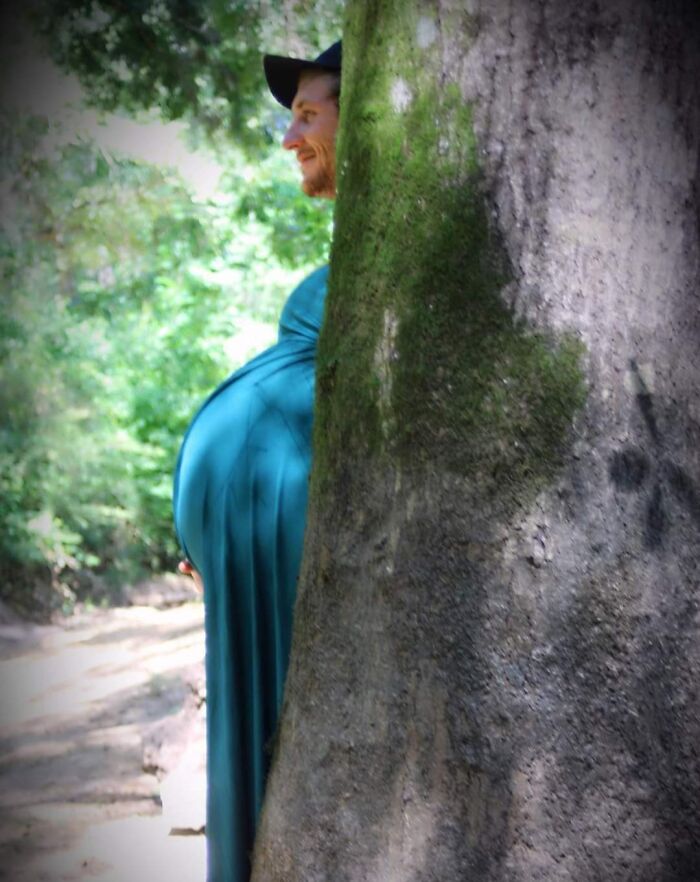 A man playfully hiding behind a tree, appearing to be pregnant, creating a funny surprise for his partner.