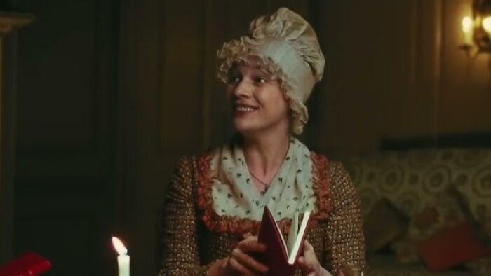 A woman in period attire smiles warmly, holding a book by candlelight.