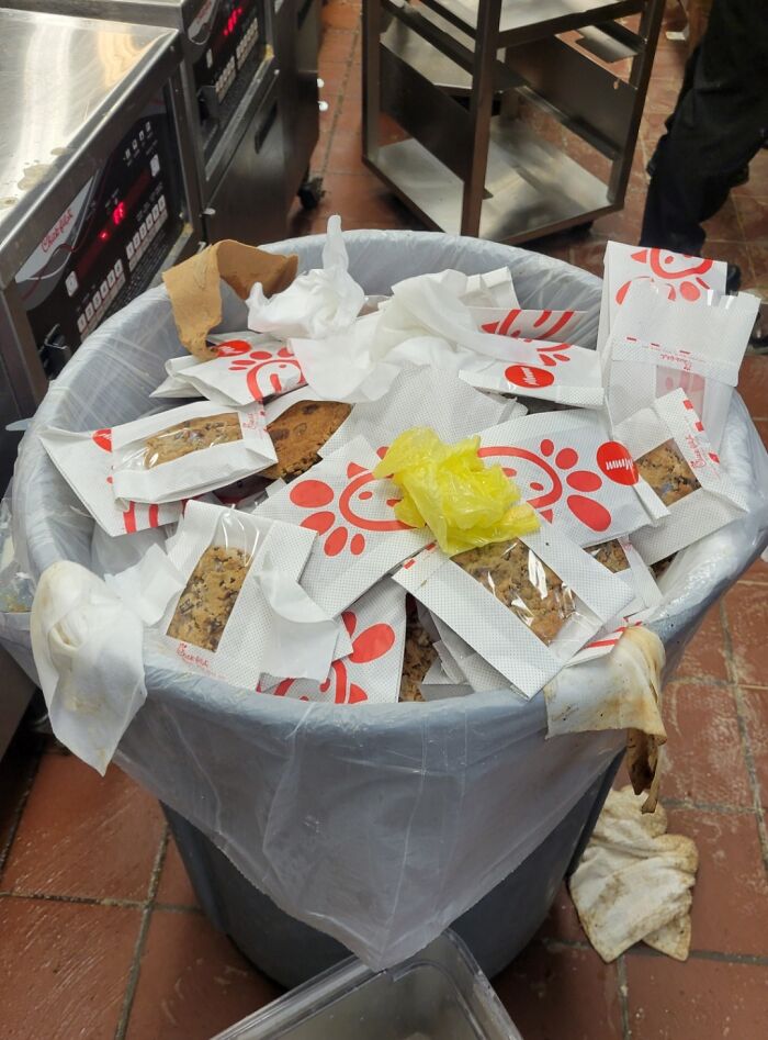 Trash can overflowing with packaged cookies in a kitchen, highlighting wasteful practices by infuriating bosses.
