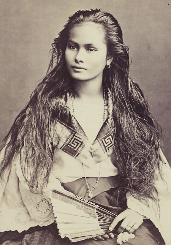 Vintage photo of a young woman with long hair wearing traditional clothing and holding a folding fan in her lap.