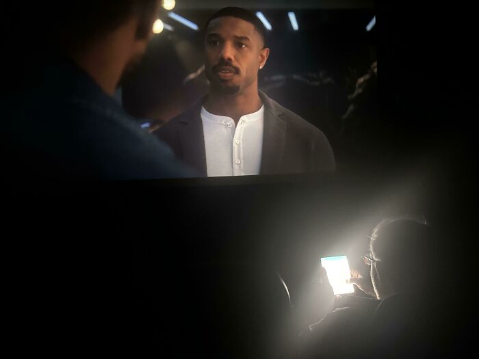 Person using phone in a dark movie theater, distracting from the film on screen, highlighting lack of basic etiquette.