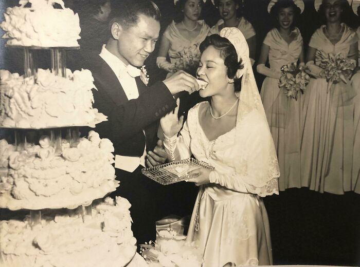 Vintage wedding photo of a groom feeding cake to his bride, surrounded by happy bridesmaids.