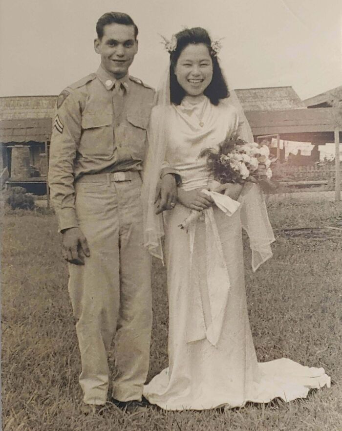 Vintage wedding photo of smiling couple, bride in a gown with flowers, groom in military uniform, standing on the grass.