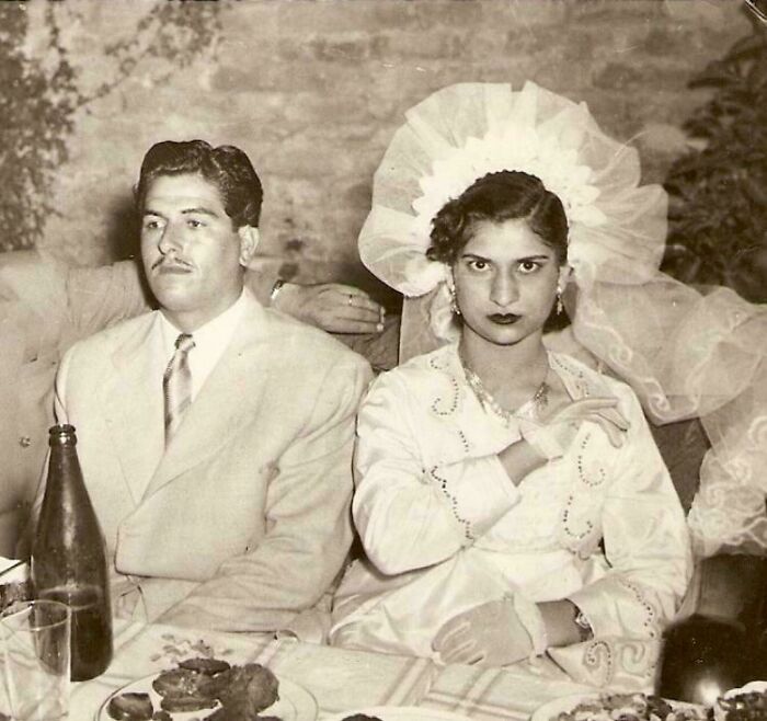 Vintage wedding couple, bride in a traditional gown with a large veil, groom wearing a classic suit at a reception table.