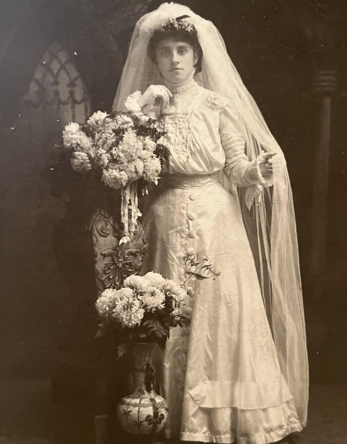 Vintage wedding photo of a bride in an elegant gown, holding a bouquet beside ornate decor.
