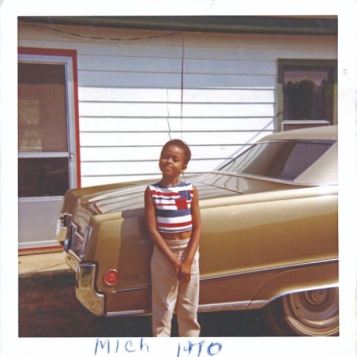 Young child stands smiling by a vintage car in front of a house, embodying a historical moment from the 1970s.