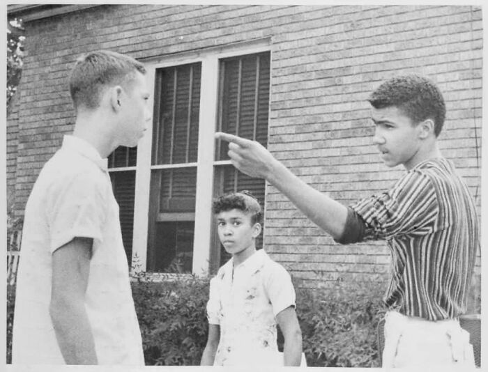 Two boys in a historical photo, one pointing and talking to another outside a brick building, capturing a past moment.
