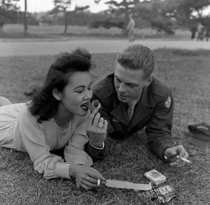 Couple in vintage setting on grass, sharing a chocolate bar and smoking, exploring historical moments.