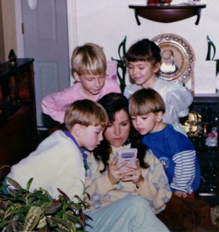 Group of children gathered around a woman playing a handheld game, capturing a moment from the past.