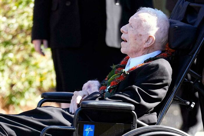 Elderly man in a wheelchair, wearing a suit and floral lei, under the sun, representing a historical moment.