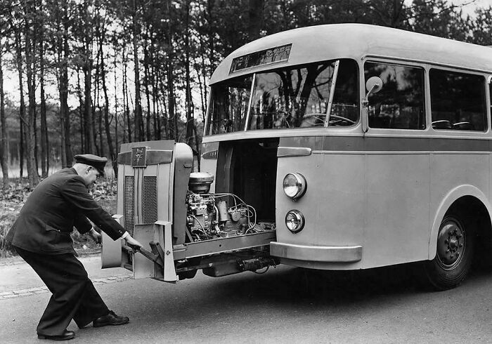 Man starting a vintage bus engine in a forest setting, showcasing historical automotive design.