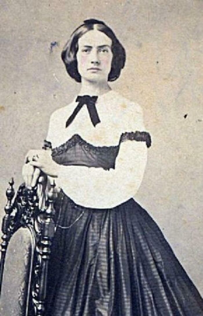 Victorian-era woman in historical dress standing beside an ornate wooden chair in an old captivating image.