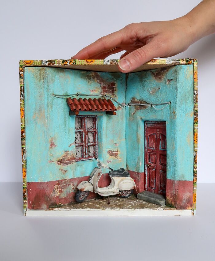 Hand holding a colorful miniature scene with a blue wall, red door, and vintage scooter, by Tamar Cohen.
