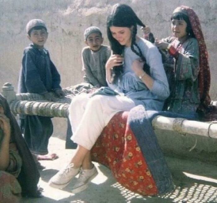 Historical photo of a woman getting her hair braided by children, seated on a woven cot, evoking a sense of the past.
