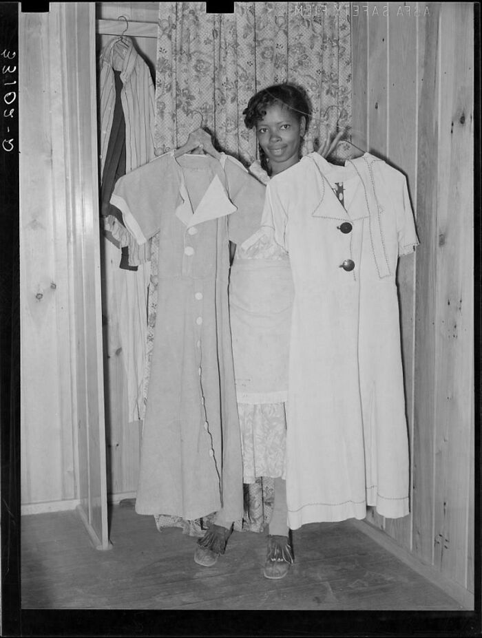 Woman showcasing vintage dresses in a wooden room, holding hangers with a smile, embodying historical fashion.