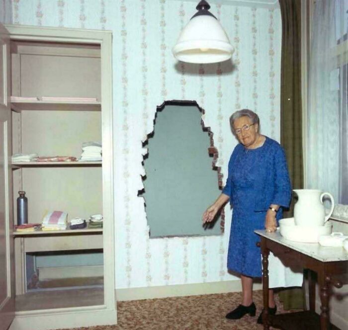 Elderly woman in blue dress points to a concealed wall opening, illustrating a fascinating historical moment.