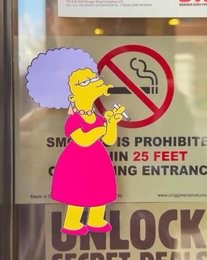 Simpsons paper cutout character standing by a no smoking sign in a real-world scene.