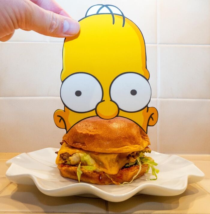 Simpsons paper cutout of Homer behind a burger, creatively merging cartoon and real-world elements.