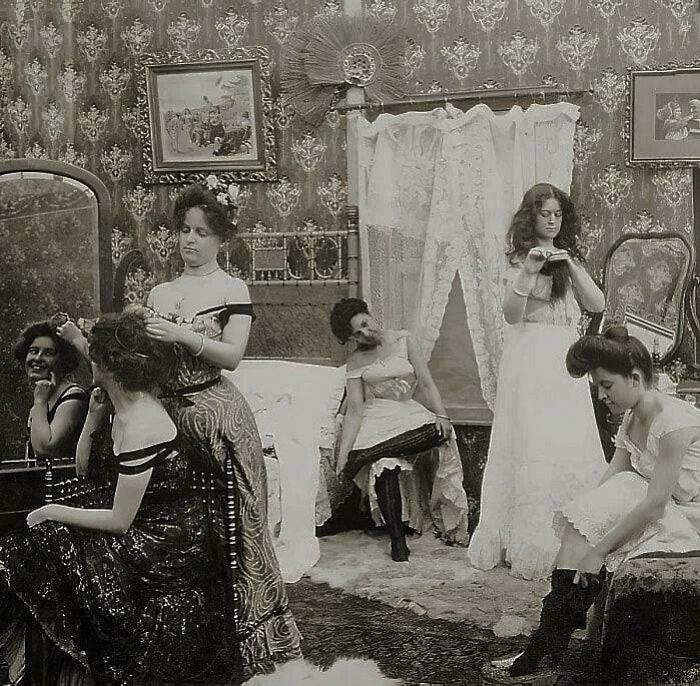 Women in vintage attire preparing hair and makeup in an ornate room, capturing a glimpse of historical fashion and beauty routines.