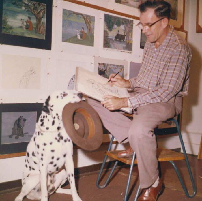 Artist drawing a Dalmatian while sitting on a chair surrounded by sketches, with a Dalmatian holding a hat nearby.
