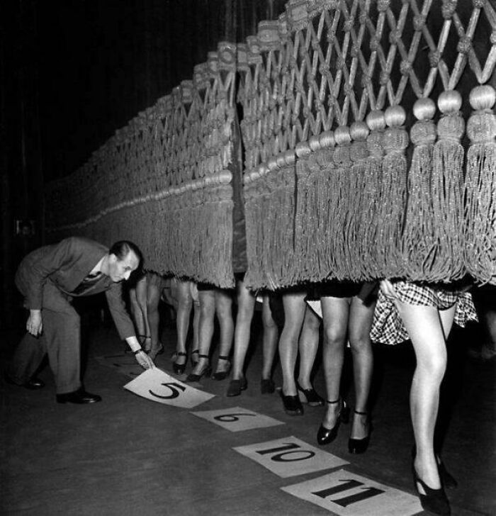 Vintage photo of dancers behind a curtain, with a man arranging numbered cards on the floor, reflecting historical era entertainment.