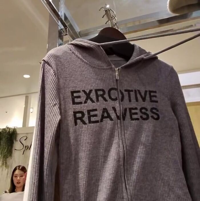 Gray hoodie with poorly translated text "EXROTIVE REAWESS" on display in a Japanese store, near a woman.