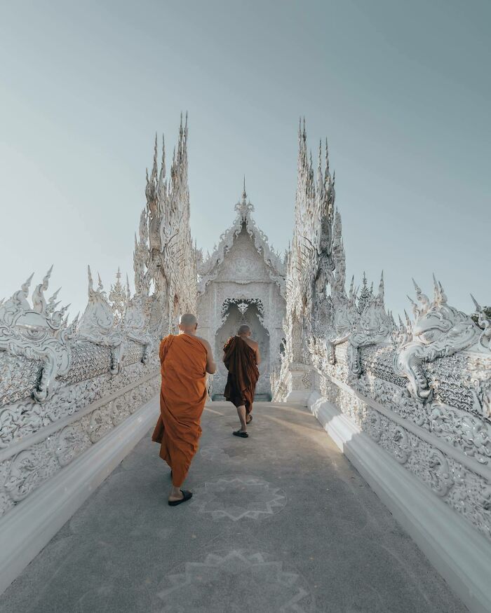 Monks in orange robes walk on an ornate white bridge, captured from an aerial perspective by a photographer.
