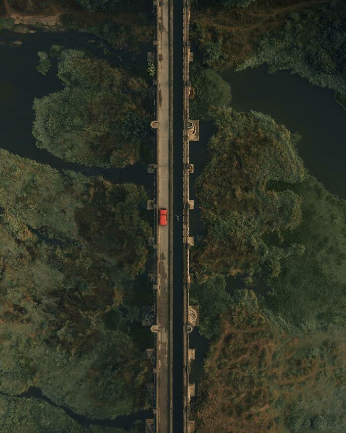 Aerial photo of a red car crossing a narrow bridge surrounded by lush greenery and water.