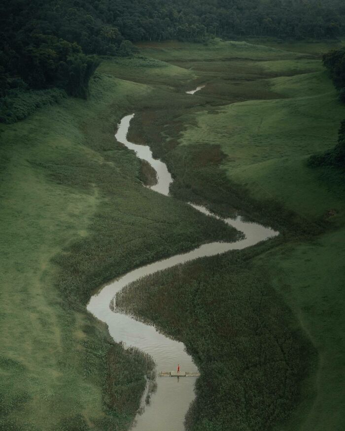 Aerial photo of a winding river flowing through green landscape with a person in a red outfit on a small raft.