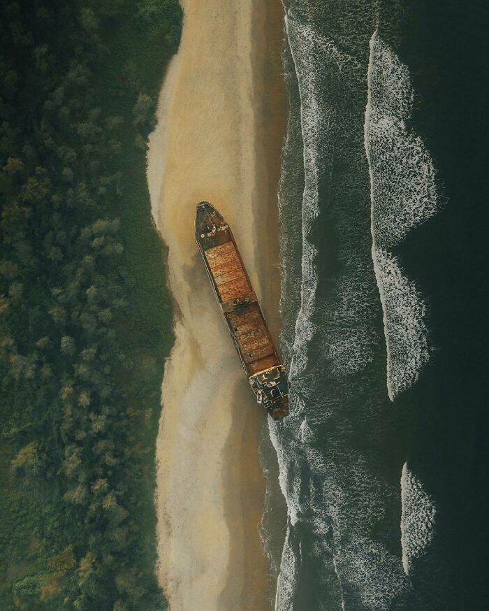 Aerial photo of a rusted ship on a sandy shore with waves approaching from the ocean.