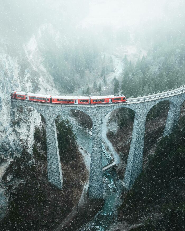 Aerial photo of a red train crossing a high stone viaduct, surrounded by misty mountains and forest.