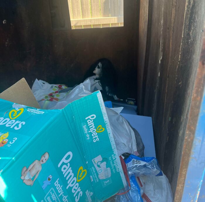 Creepy doll with black hair hidden in a dark corner behind a Pampers box and plastic bags.