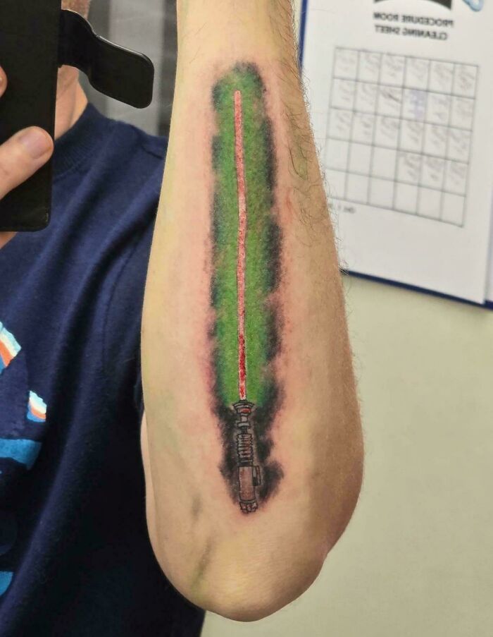 Tattoo fail of a red lightsaber with smudged green and black ink on a person's inner forearm, showing poor design and e*******n.