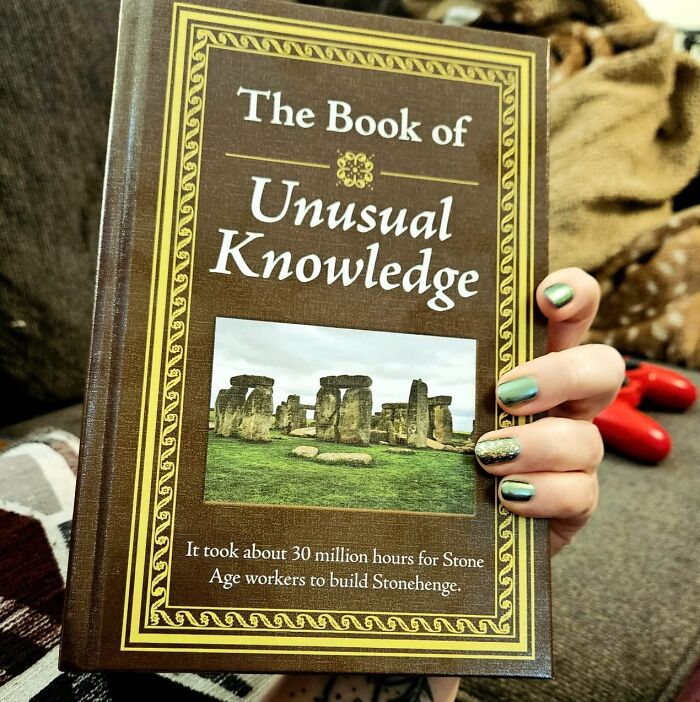 A hand holding "The Book of Unusual Knowledge," featuring Stonehenge on the cover.
