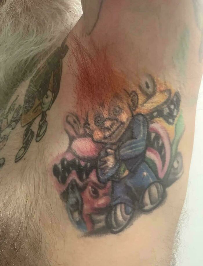 Tattoo fail of a distorted cartoon character with mismatched colors and unclear design on a hairy arm.