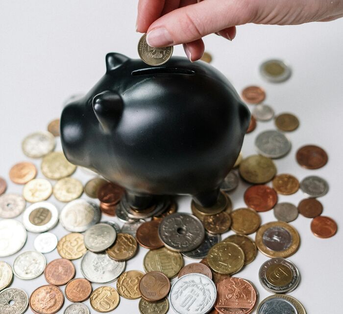 Hand placing coin into black piggy bank surrounded by scattered coins, symbolizing lottery winnings and money management.
