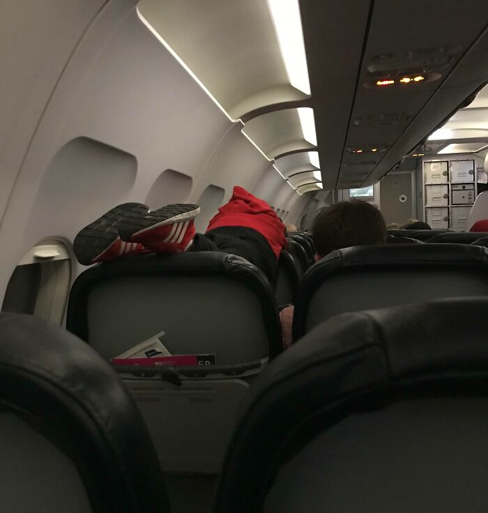 Passenger in red shirt lying across airplane seats, shoes on headrest, displaying poor etiquette.