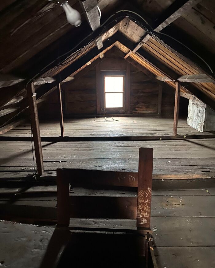 Ático oscuro y abandonado en casa nueva con vigas de madera y ventana iluminada, que genera sensación de sorpresa y miedo.