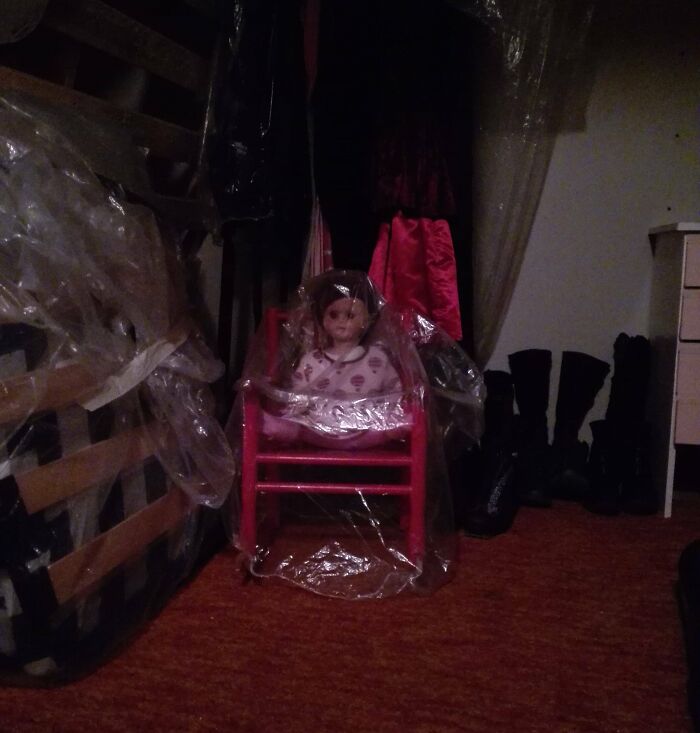 Creepy doll wrapped in plastic sitting on a red chair in a dimly lit room, found hidden in a home after moving in.