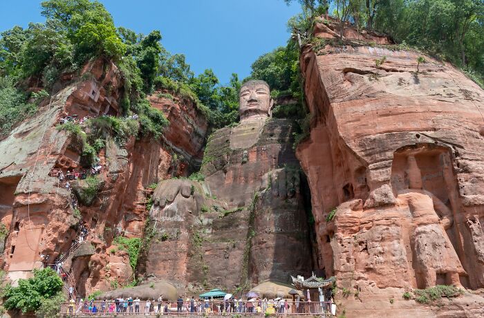 Giant stone Buddha sculpture carved into a red cliff, surrounded by lush greenery, exemplifying architectural wonders.