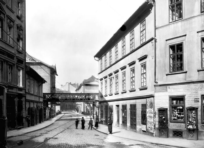 Street scene in Europe 100 years ago showing cobblestone roads, historic buildings, and pedestrians in early 20th century attire.