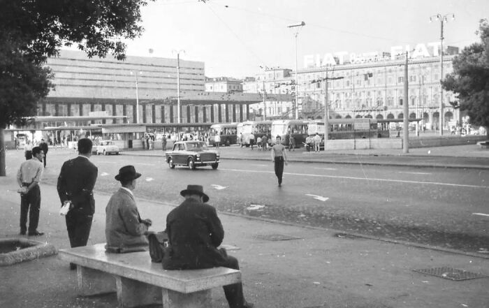 Black and white photo of daily life in Europe 100 years ago showing people waiting near a street with vintage cars and buildings.