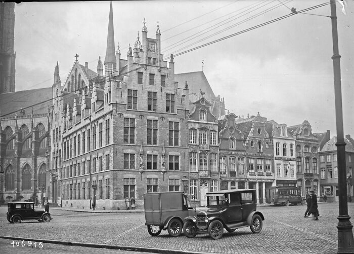 Historic street scene in Europe featuring early 20th-century buildings and vintage cars, illustrating life 100 years ago.