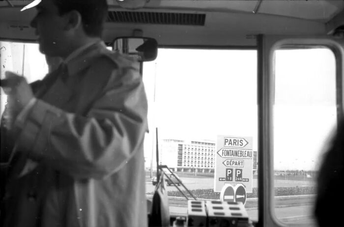 Man in coat inside vehicle near road sign pointing to Paris and Fontainebleau, showing life in Europe 100 years ago.