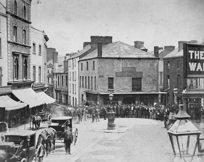 Crowded European street scene from 100 years ago showing life with horse-drawn carriages and historic buildings.