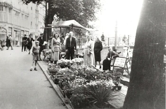Black and white photo showing a European street flower market with people and children, depicting life in Europe 100 years ago.
