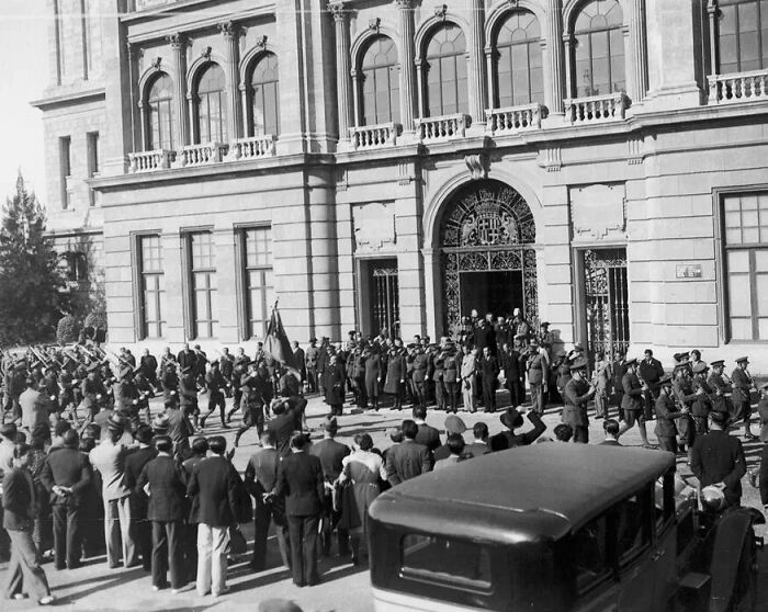 Crowd gathered outside historic European building with soldiers and vintage car, life in Europe 100 years ago.